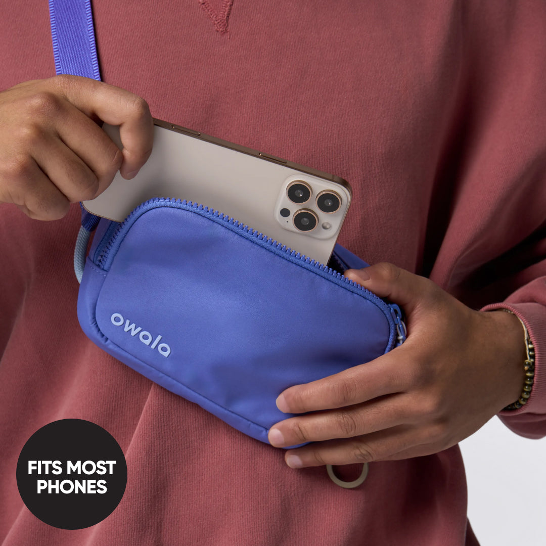 Owala Daily Crossbody in Al Fresco, peach belt bag with matching strap worn over the shoulder of a woman in a white t-shirt on a rooftop overlooking a city skyline